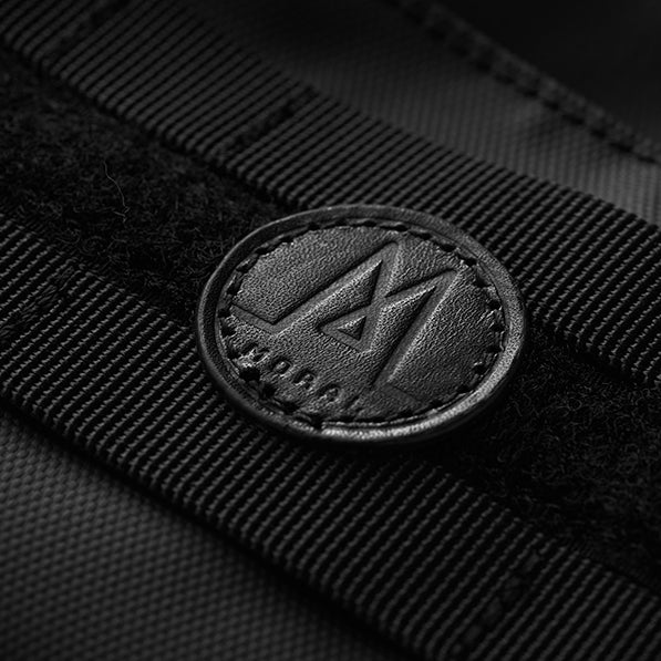 Moral NIGHTHAWKS MILITARY DUO MINI 軍事風側背包品牌標誌特寫,展現細緻的皮革圓形Logo設計。精緻縫線與軍事風織帶,突顯質感與識別度。